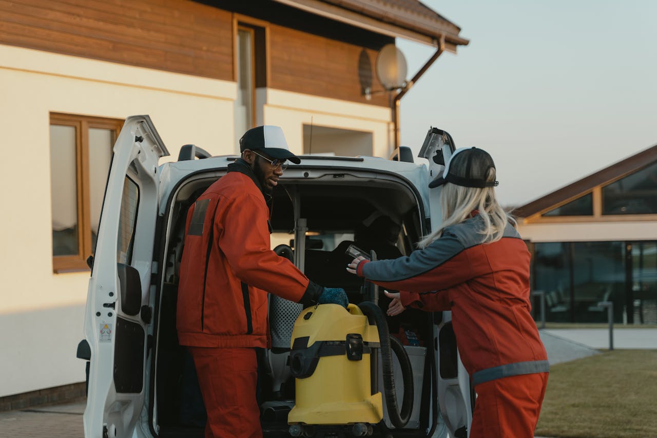 about-01 Two cleaners in red uniforms load a vacuum into a white van outdoors.